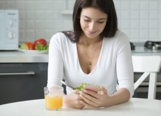 Image of a woman sitting in a kitchen using a smartphone to record drinking an orange juice into her diet apps