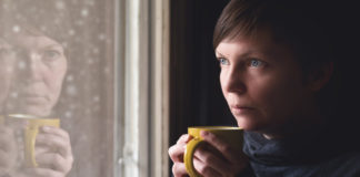 Lonelsome woman drinking cup of coffee by the window of her living room, looking out at snow falling with a sad look on her face. Selective focus with shallow depth of field.