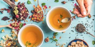 Health benefits of herbal tea Two cups of healthy herbal tea with mint, cinnamon, dried rose and camomile flowers in spoons and man's hand holding spoon of honey, blue background, top view
