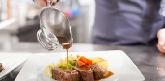 Chef plating up food in a restaurant pouring a gravy or sauce over the meat before serving it to the customer, close up view of his hand and the gravy boat