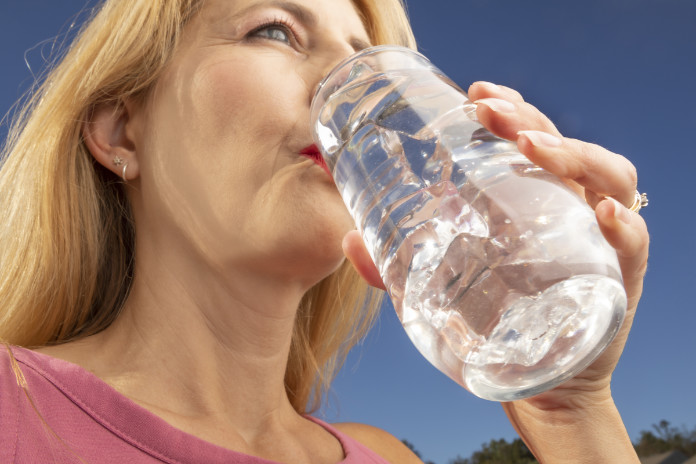 A close up of a 47 year old woman drinking a glass of ice water outdoors.