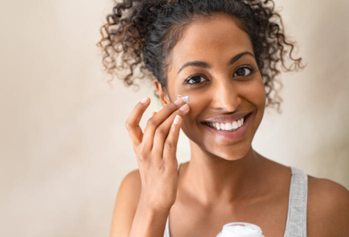 Smiling african girl with curly hair applying facial moisturizer while holding jar and looking at camera. Portrait of young black woman applying cream on her face isolated on beige background. Close up of happy attractive beauty woman caring of her skin standing on light brown wall with copy space.