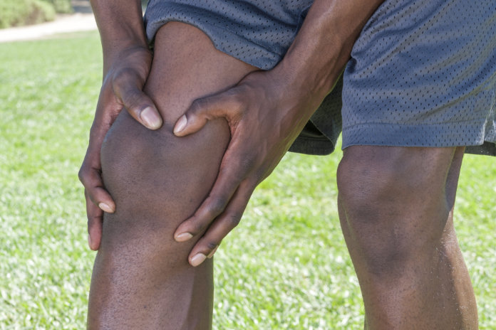 Closeup of knee and leg of lean African American male athlete clutching injured knee with fingers around the patella on green lawn outdoors