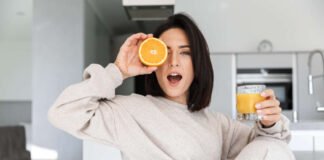 Vitamin C foods – smiling woman 30s drinking orange juice while resting in bright modern room