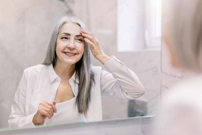 Pretty senior woman with long gray hair, wearing white shirt, looking at her face in the bathroom mirror, and applying anti-wrinkle cream or cosmetic