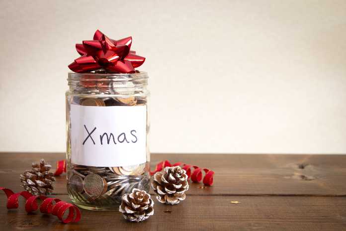 A jar of coins on a wooden table with Christmas decorations and a hand written sign, on a white background.