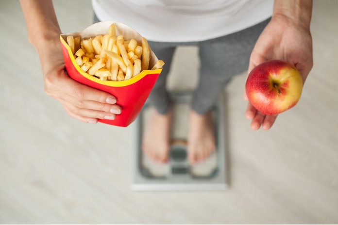 A man holding an apple and some fries while stood on scales. 