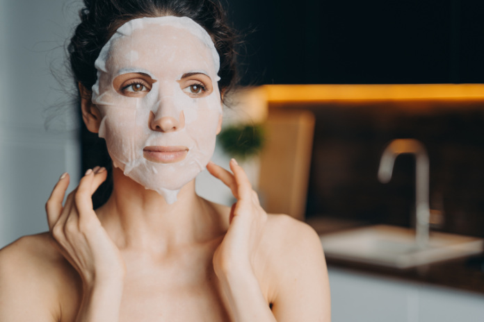 Female with naked shoulders uses rejuvenation facial sheet mask. Skin treatment at home, skincare